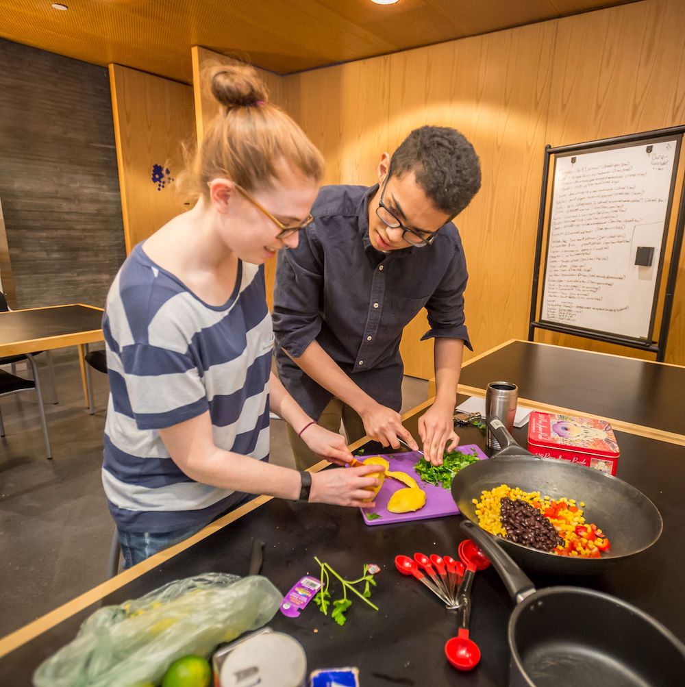 Two students place food in pans as they cook together in a dormitory community kitchen.