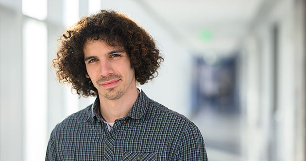 Graduate student, Eli Sanchez standing in a out of focus naturally lit hallway, in the left side of the photograph, MIT