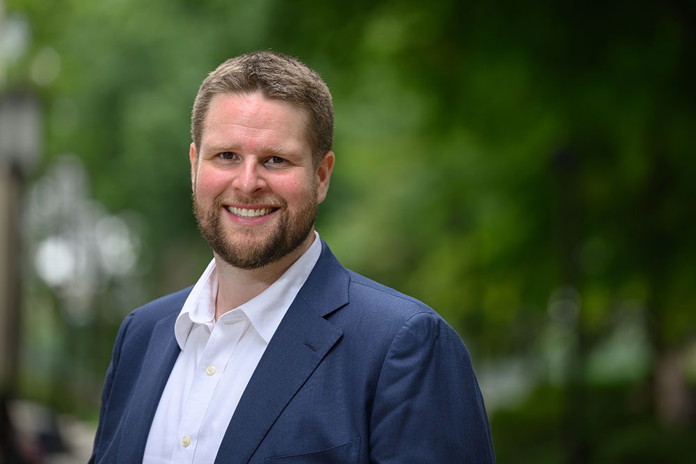 Headshot of faculty member Ethan Peterson, outdoors against a green trees, MIT