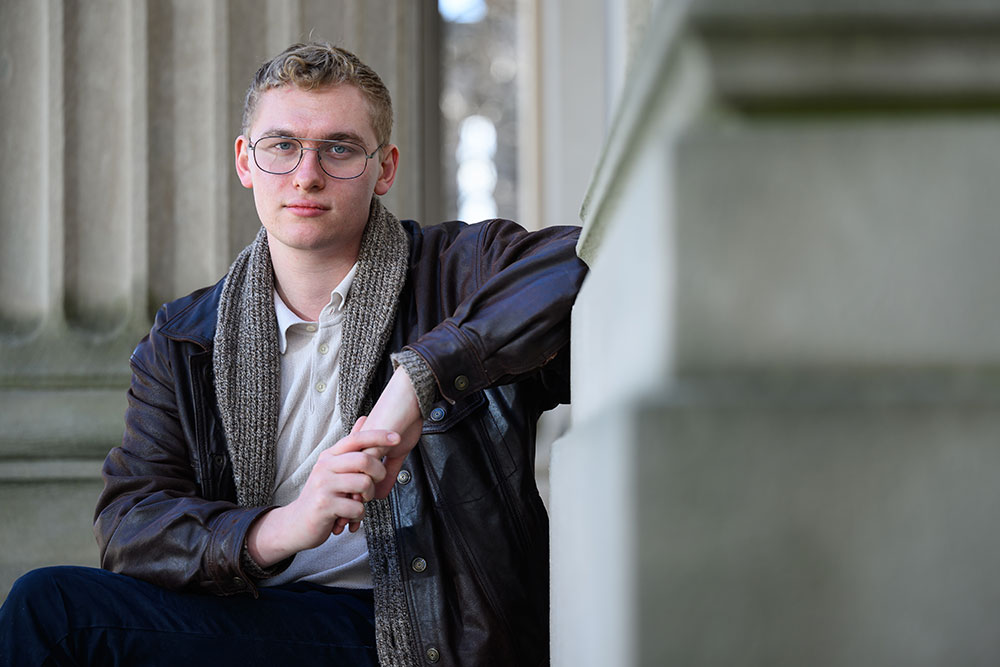 Graduate student, Liam Jines, seated outdoors with concrete architectural columns in the background and to the right