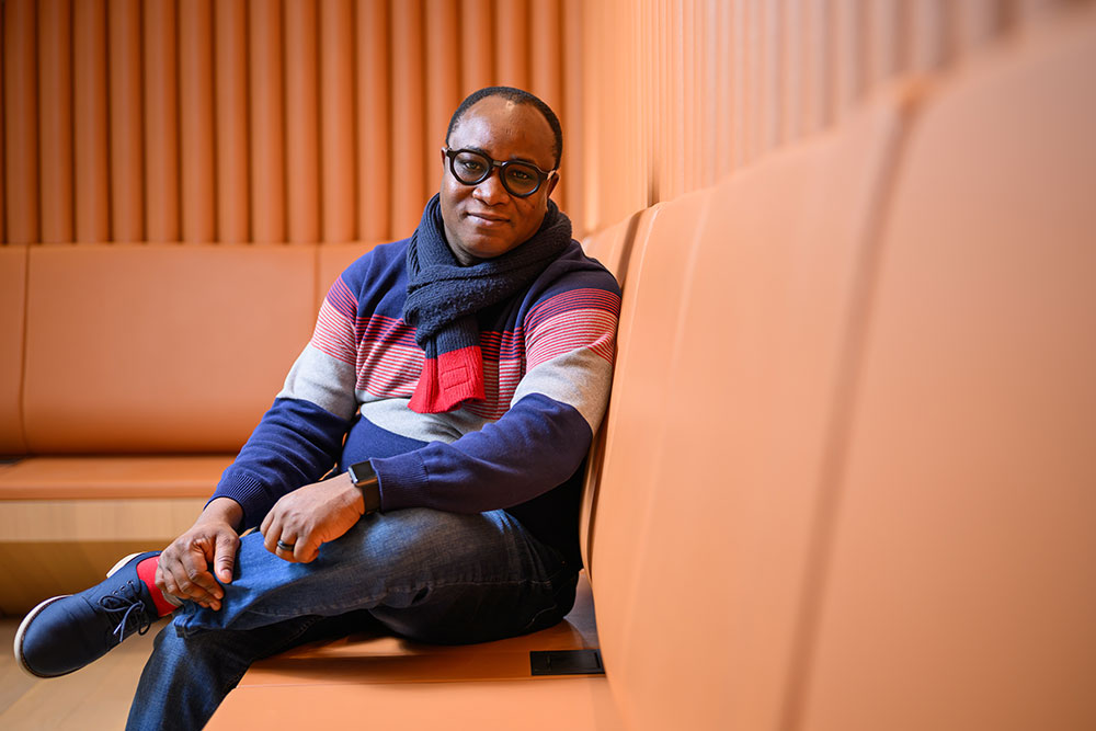 Male Professor Ericmoore Jossou, seated leaning on his left arm, right leg crossed over left on an orange sofa indoors, MIT